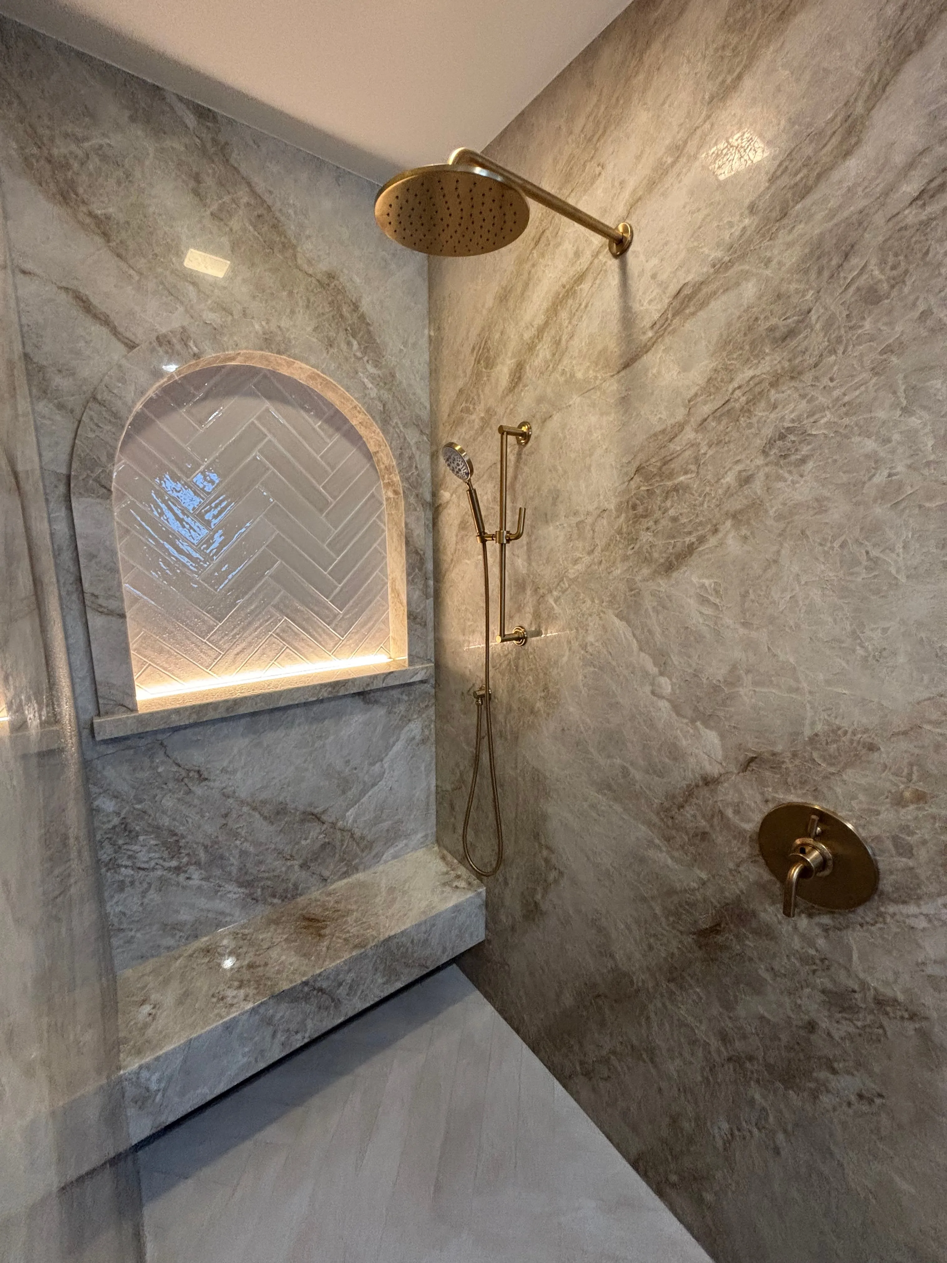 Marble walk-in shower with brushed brass rain head, hand shower, and lit niche