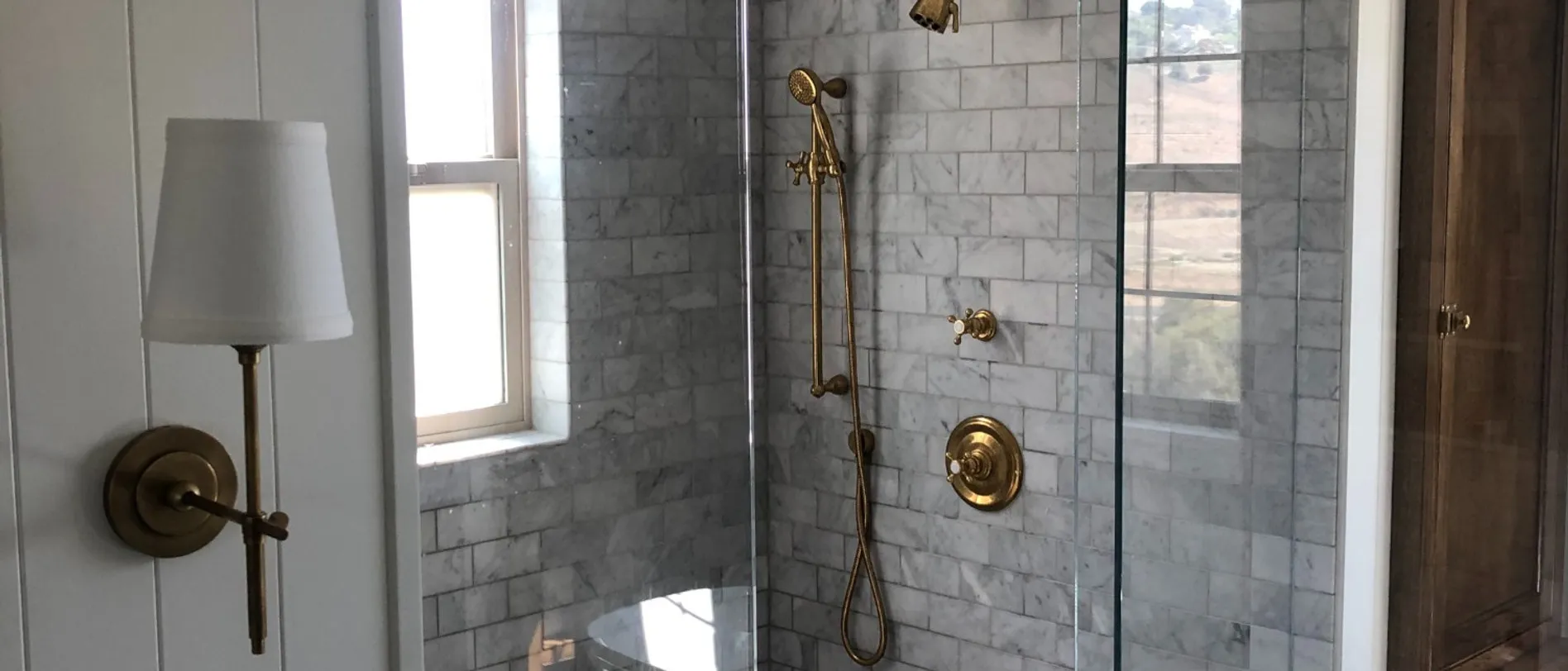 Walk-in shower in marble subway tile with an unlacquered brass shower set and hand shower