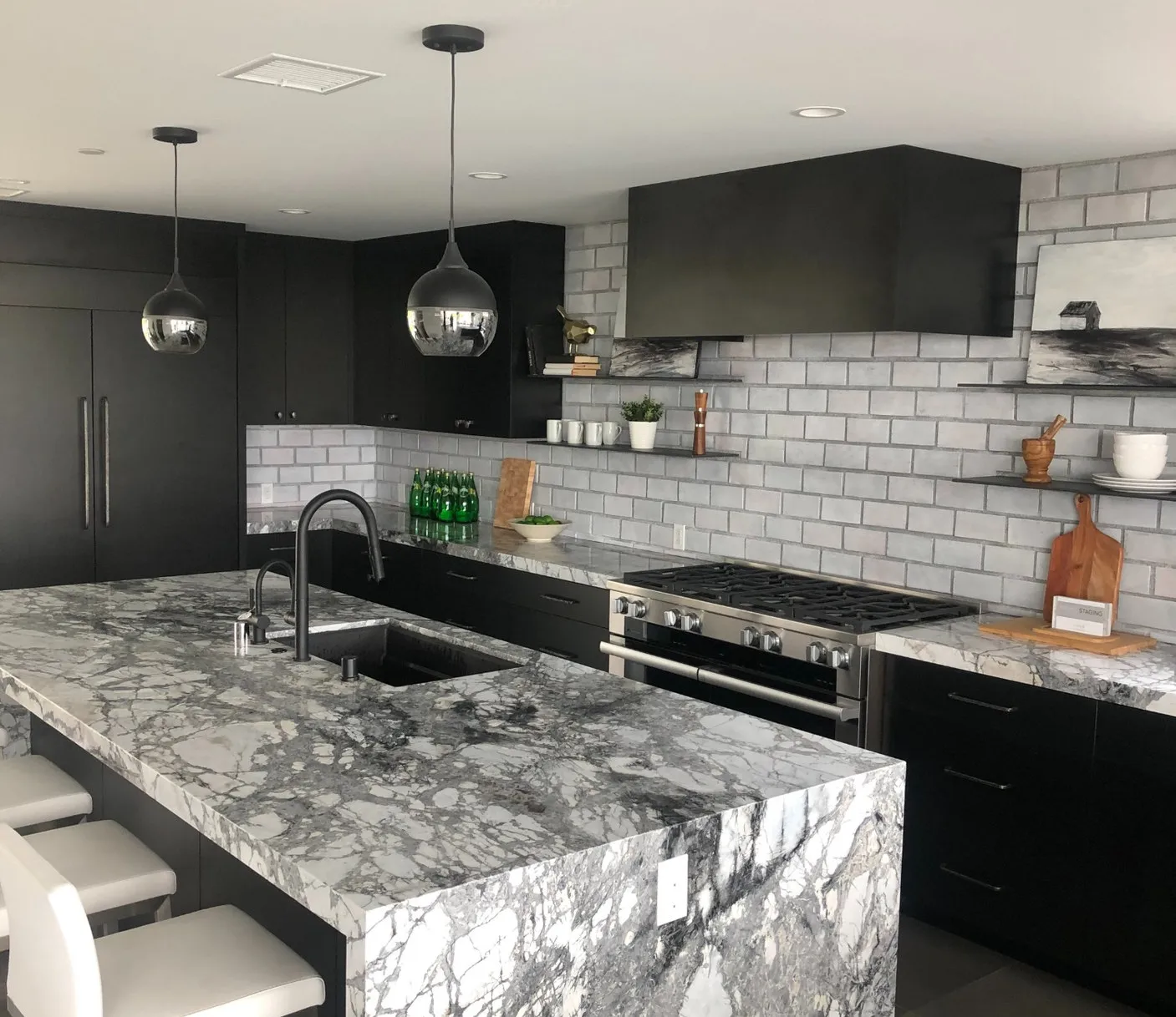 Black-cabinet kitchen with a veined marble waterfall island, chrome pendants and a pro gas range