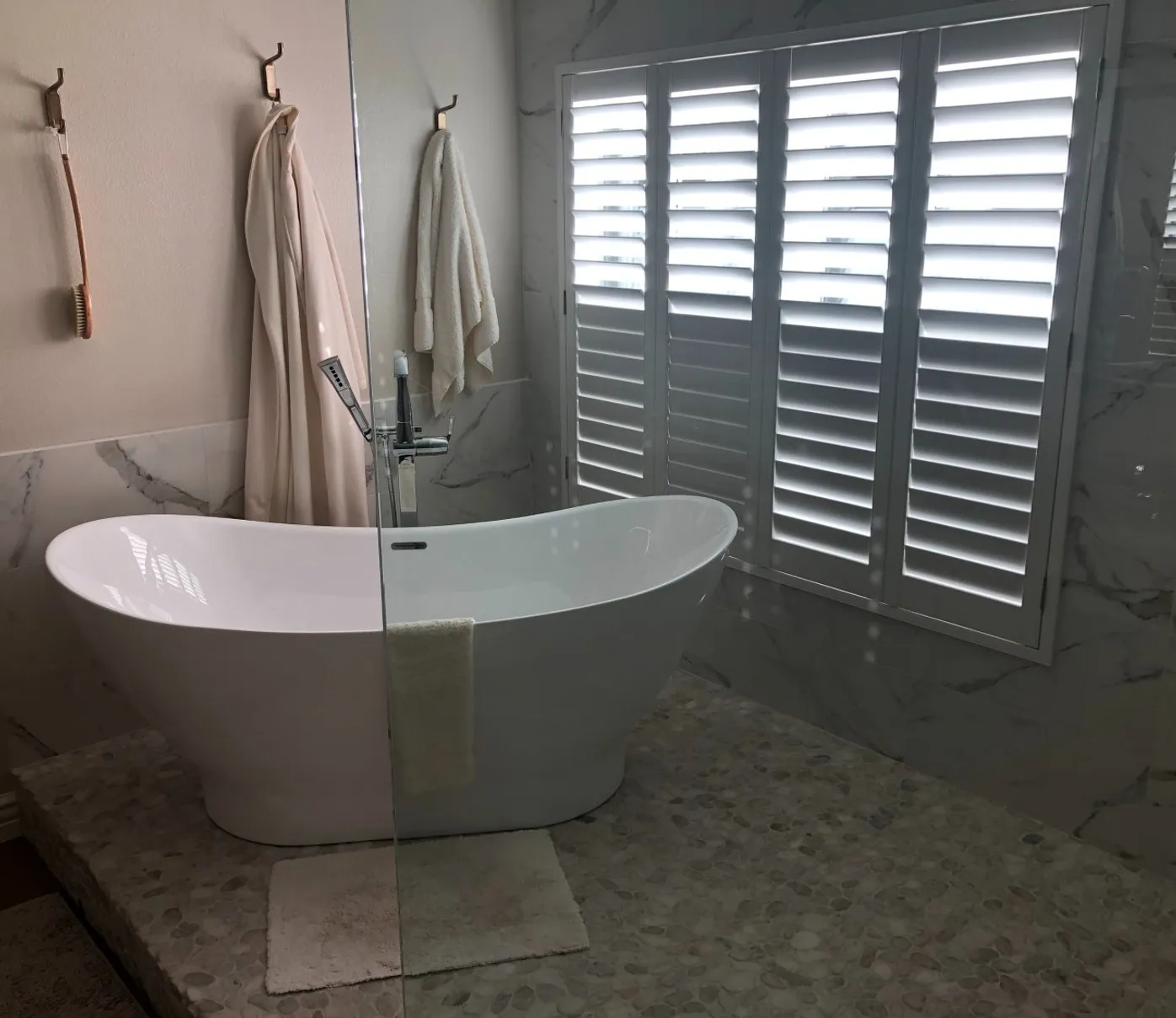 Freestanding slipper tub on a pebble-tiled platform against marble walls with plantation shutters